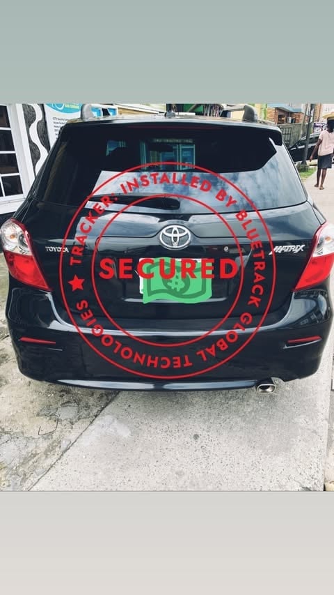 Black Toyota hatchback with red circular security seal and green logo on rear window, parked on street